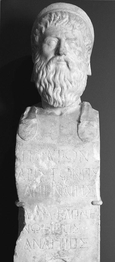 A bust of an ancient figure. The figure is wearing a head covering and has short wavy hair, a mustache, and a beard. Greek characters can be seen chiseled in the marble below the bust.