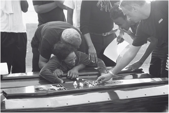 Figure 5. A woman consoled by a man mourns by a coffin. Another man places flowers under a photograph taped on the coffin.
