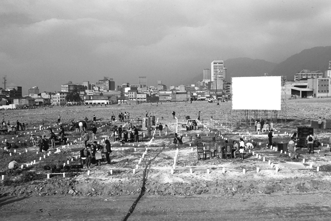 Figure 2.2. A dirt field with seating areas and groups of people standing near a large white screen and between rows of white candles on the ground. City buildings and hills are visible in the distance.