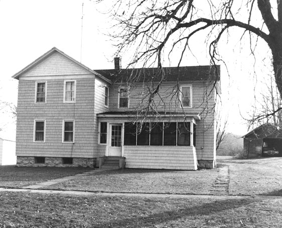 Figure 10 A two-story, wood-shingle-covered, L-shaped house has an enclosed single-story porch at the front entrance. The plain-looking house is surrounded by a grass yard with no plantings.