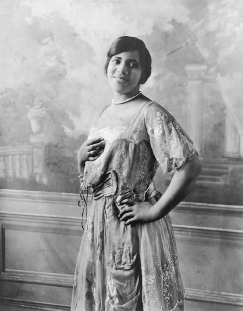 A smiling African American woman wearing a string of pearls stands in concert dress against a mural.