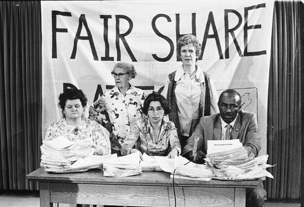 Figure 50. Four women and one man behind a desk piled high with signed petitions. Behind them is a sign that says Fair Share.