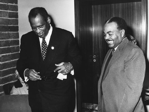 Two Black men wearing suits are laughing and smiling with each other while walking.