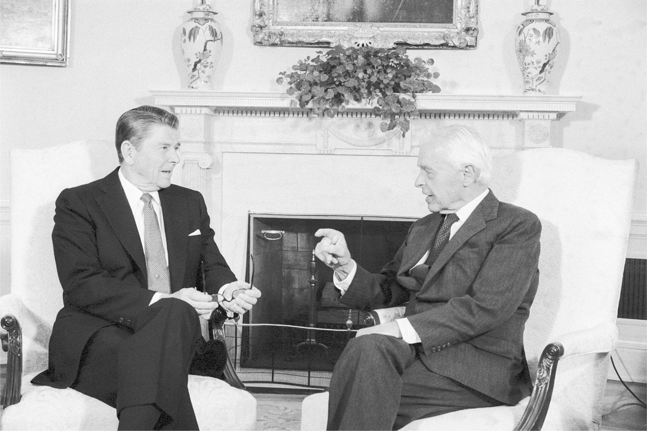 Reagan and Nitze are conversing while sitting on chairs in front of a fireplace. Both men are wearing suits and ties. Nitze is gesturing to Reagan with his right hand.