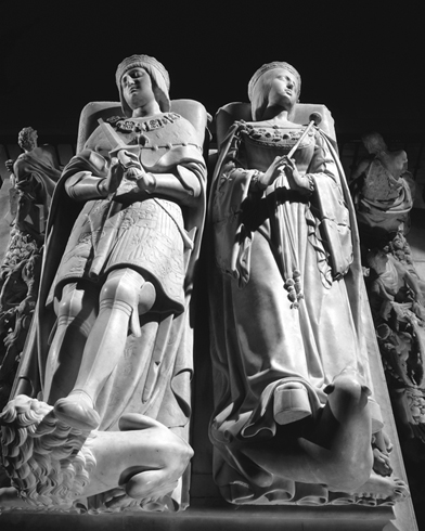 Seen from above, a carved alabaster tomb featuring a kingly figure holding a sword and with his feet on a small lion, while on the right, a queenly figure reclines with her feet on a dog, with the couple looking away from each other.