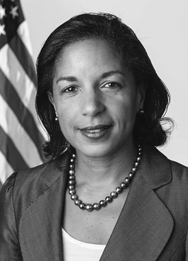 A portrait of a woman looking directly at the camera. She has dark shoulder-length hair and is wearing a blazer over a light-colored shirt with a necklace and earrings. The US flag can be seen in the background over her right shoulder.