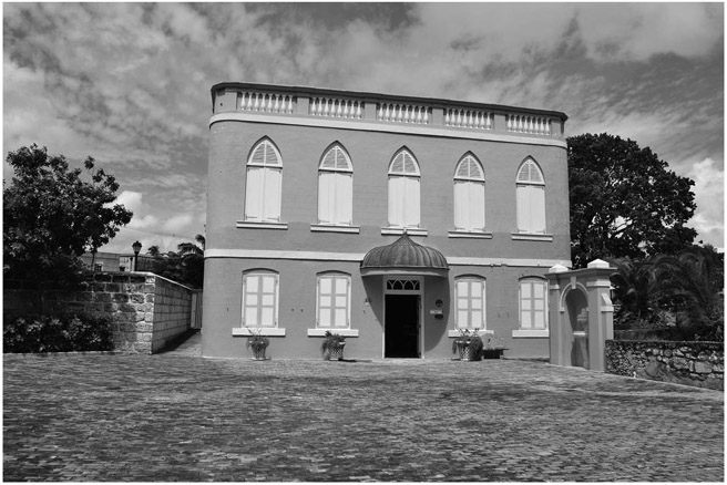 Figure 9.1: A building with four windows and a door on the ground floor and five windows on the first floor. On the left of the building is a wall with bushes in front of it and a tree behind it. To the right of the building are a gate and a low wall.