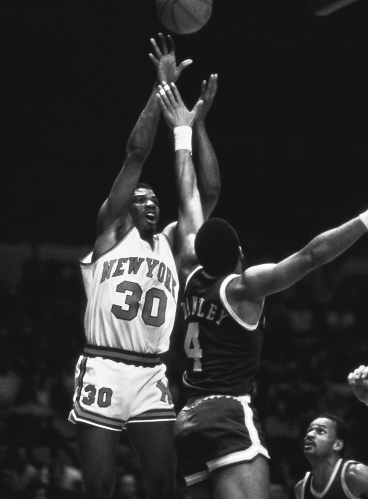 An open-mouthed King jumps and lifts the ball with arms fully raised as Dantley also jumps and reaches for the ball.
