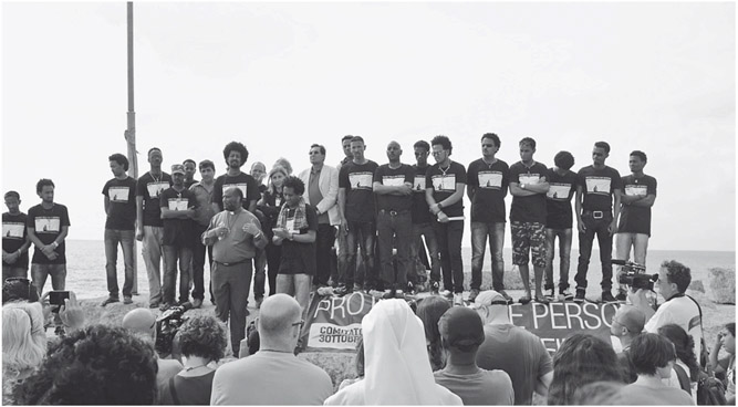 Figure 14. A group of about twenty people, most of them wearing identical T-shirts stand on an elevated construction in front of a crowd of people gathered to watch and listen to them.