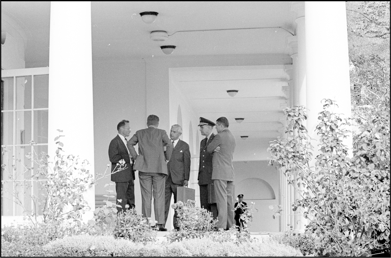President Kennedy is briefed by four members of the Executive Committee of the National Security Council in a colonnade outside the White House. All four look very serious. Kennedy’s back is to the camera. Nitze is holding a briefcase.