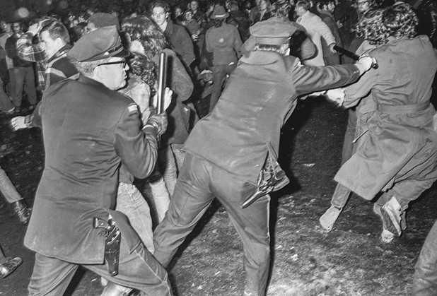 Figure 35. Two policemen charge into crowd of protestors. The lead one is hitting a young protestor who is wearing a long coat. The other is pushing a protestor who is hunched over trying to protect himself.