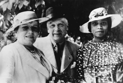 Three figures stand outside underneath a tree and look directly at the camera. On the left is an older white woman in a white dress suit holding a bouquet, in the center is an older white man, and on the right is a young Black woman wearing a print dress and a straw hat.