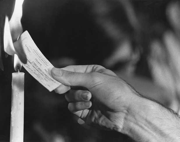 Figure 12. A man’s hand holds a draft card up to the flame of a church candle. The draft card is burning, its flame extending the flame of the candle.