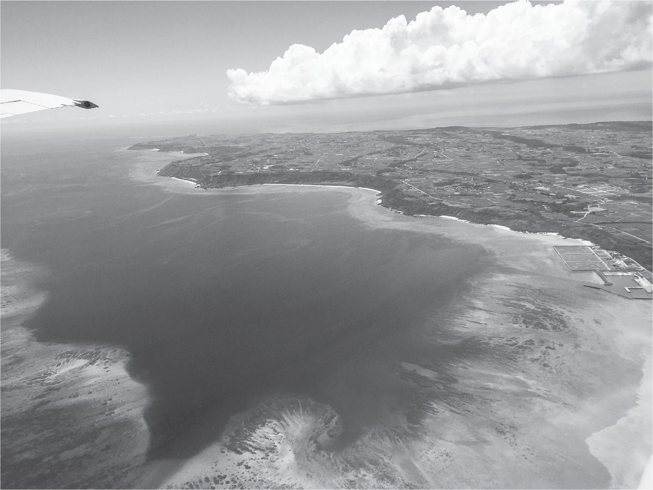 An aerial view shows the clear blue, sweeping shorelines of Miyakojima. A string of clouds floats above the island, and an airplane wing barely juts into view.