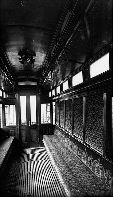 Inside a streetcar showing wooden flooring, two lines of benches, heavy shades drawn over the main windows, small windows at the top, and a door.
