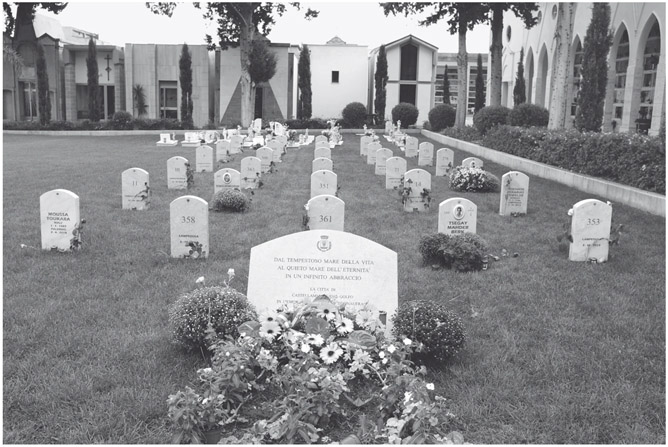 Figure 9. A large memorial plaque with Italian text stands in front of a cemetery of thirty numbered graves with similar headstones on a green lawn. There is a small flower bed in the front. Three headstones have photos, and two just names.