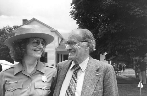 Figure 13 A curly-haired women in a ranger uniform shirt and flat-brim ranger hat poses alongside a white-haired man in jacket and striped tie. They are smiling broadly at each other. Small groups of people stand in the background in the yard of the restored house.