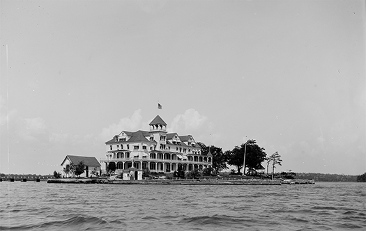 A big house with an American flag on the roof sitting on an island in a river, with the shore visible behind it.