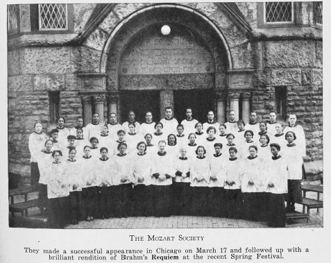 A group of approximately fifty young men and women stand in three rows wearing white robes outside of a building.