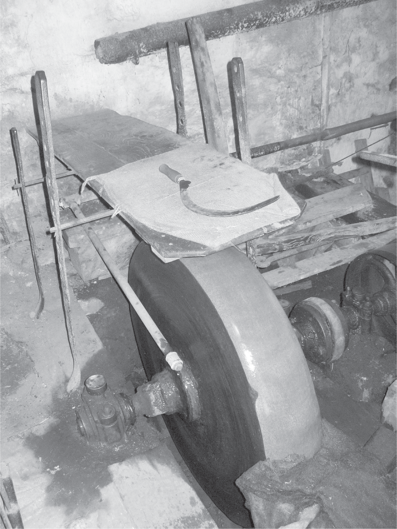 Figure 20: A body-length plank grindstone photograph is shown from a three-quarter angle. On the left side of the photograph, there is a ladder one ascends to access the grindstone. The plank is positioned from back to front, where the grinder would lie with feet in the back, head and face in the forefront of the photograph just above the enormous grinding wheel.
