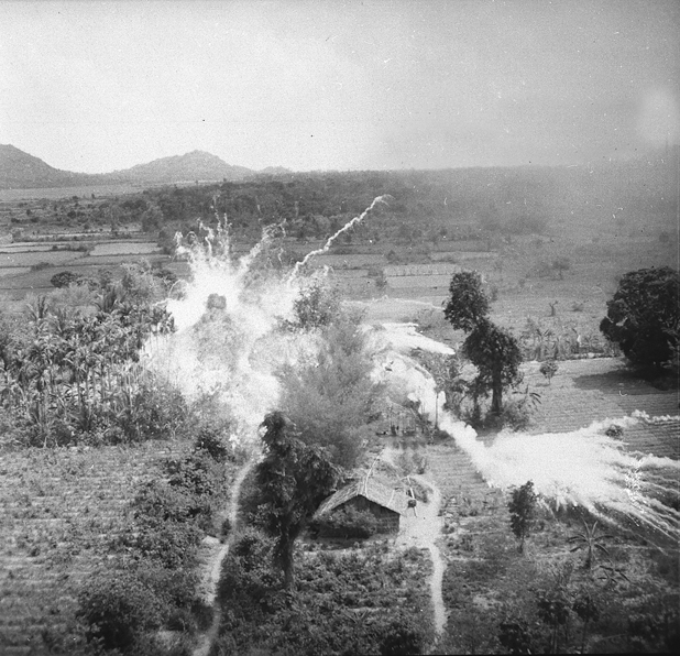 Figure 14. A fiery napalm explosion destroys a cluster of houses in Vietnam, the surrounding countryside is green, the napalm bright orange with tendrils of smoke snaking off in several directions.