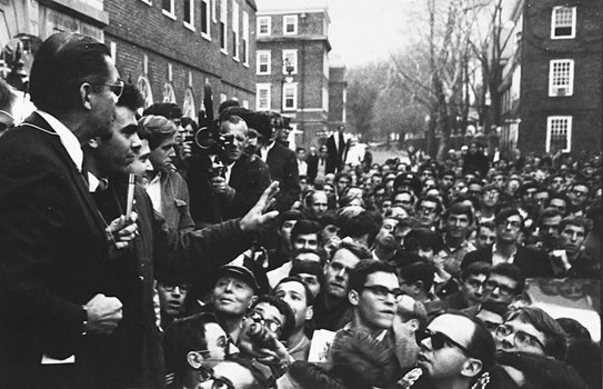 Figure 8. Secretary of Defense McNamara is up on a car hood holding a microphone. Standing next to him is a young man (me) speaking to crowd of hundreds of protesting students clogging a narrow Cambridge Street outside of Harvard’s Quincy House.