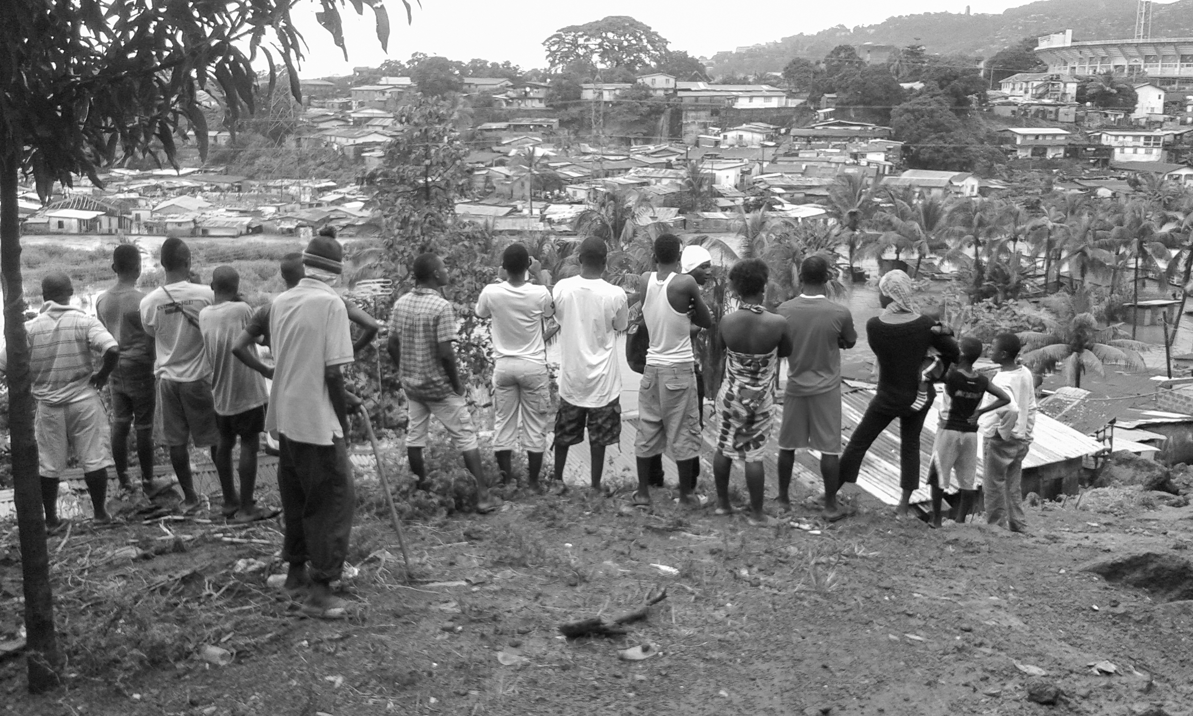 Figure 2. Many people stand on a hill with their backs to the camera, looking at the land with houses and trees below.