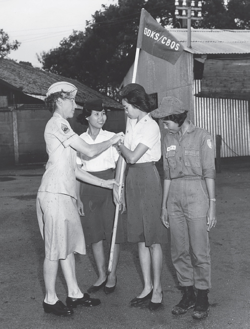 An American WAC helps a Vietnamese WAC trainee learn to hold a flag in formation on August 8, 1966. Two fellow Vietnamese trainees stand to either side smiling.