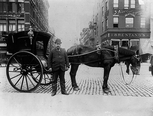 A horse-drawn cab on a cobblestone street. The driver stands in front of the cab and the horse. City buildings are visible in the background.