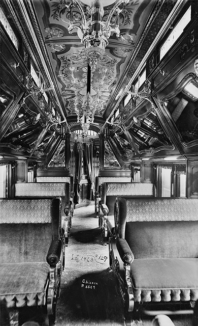 Inside a luxurious railroad passenger car with intricate woodwork in the ceiling and mirrors above plush bench seats.