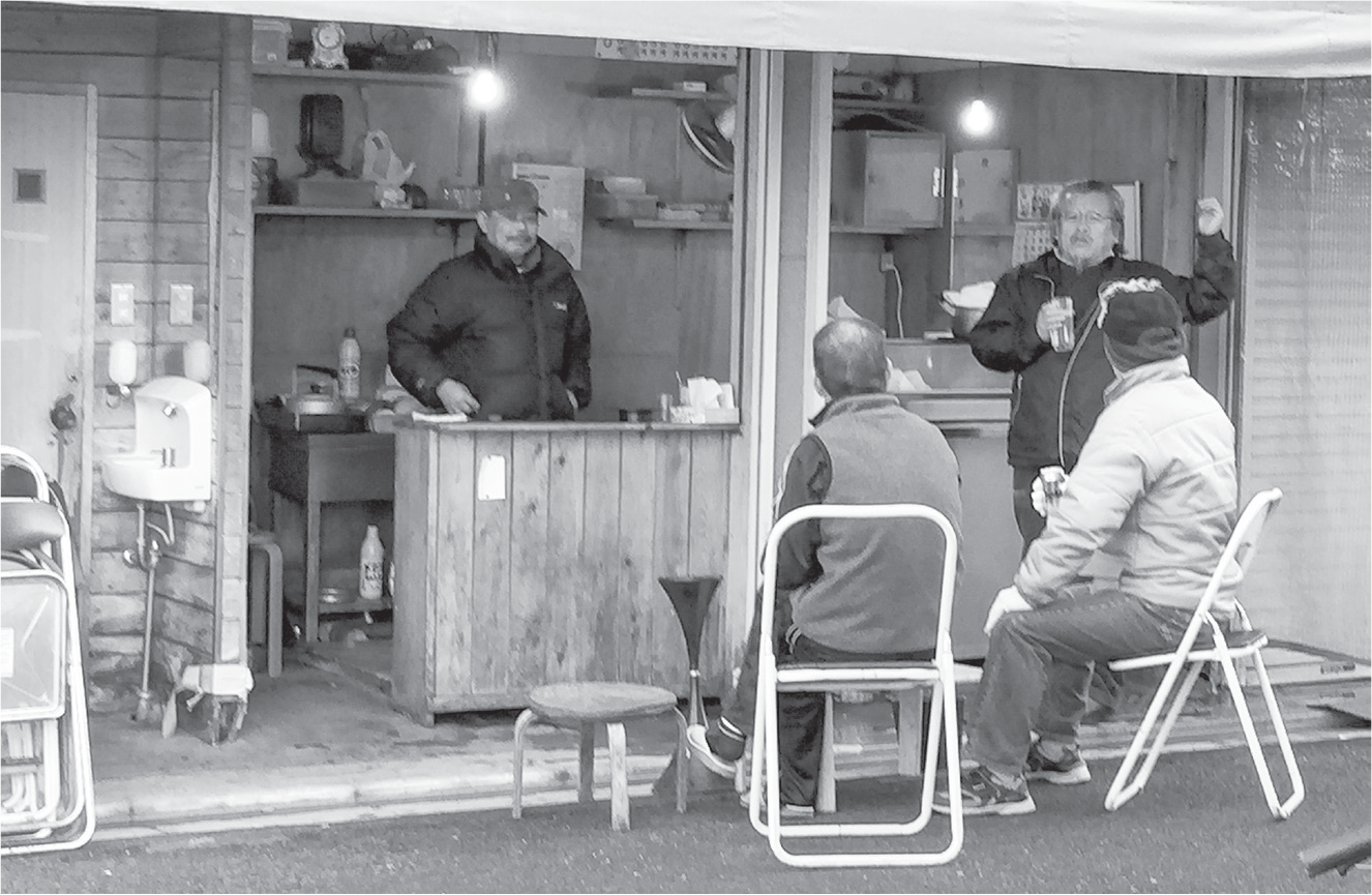A sushi chef stands behind a wooden counter, as three men stand and sit on plastic chairs, talking on the street. The chef is wearing a jacket to stay warm. A ceramic sink adjoins the counter.