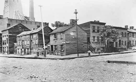 Figure 3.3. Nine shabby, two- and three-story, wooden houses, line a cobblestone street near the South Brooklyn waterfront, in an old, lantern-slide photograph. A factory or grain silo rises behind these workers’ homes.
