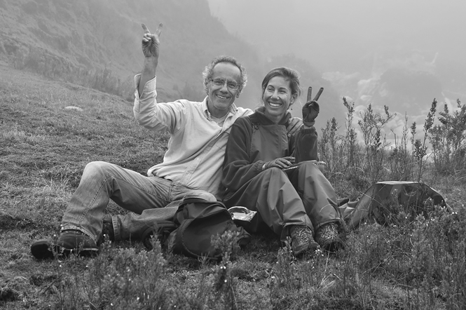 Patricia Burrowes and Ignacio De la Riva sit next to each other on the ground smiling, each with a hand up making a V with their fingers.