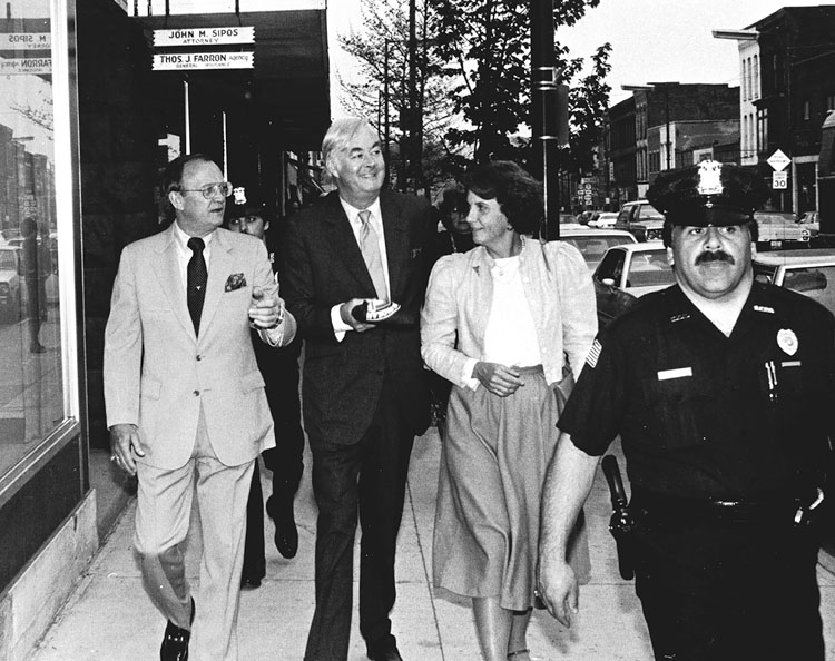 Figure 6 A smiling white-haired man in a dark suit is flanked by a man and a woman, with police officers in front and behind, as he walks down the sidewalk.