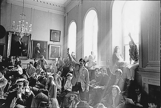 Figure 21. An ornate room jammed with young students. The light filters through three large windows. In the middle is a young man, hair falling over forehead, with a dark mustache, his hand raised. A glass chandelier hangs from the high ceiling. On the walls are old paintings of men.