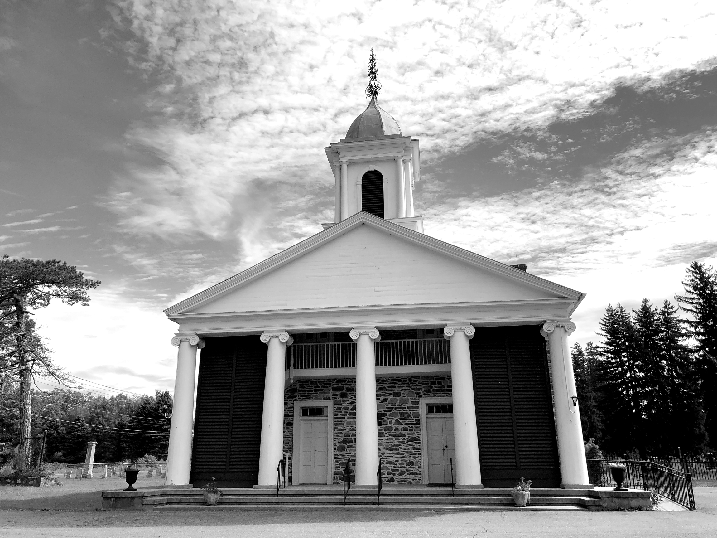 A church with a white portico with five white columns. A few steps lead to the portico. The building has two white doors as entrances on the first floor. The bell tower peaks out over the portico. A graveyard is visible on the sides of the church.
