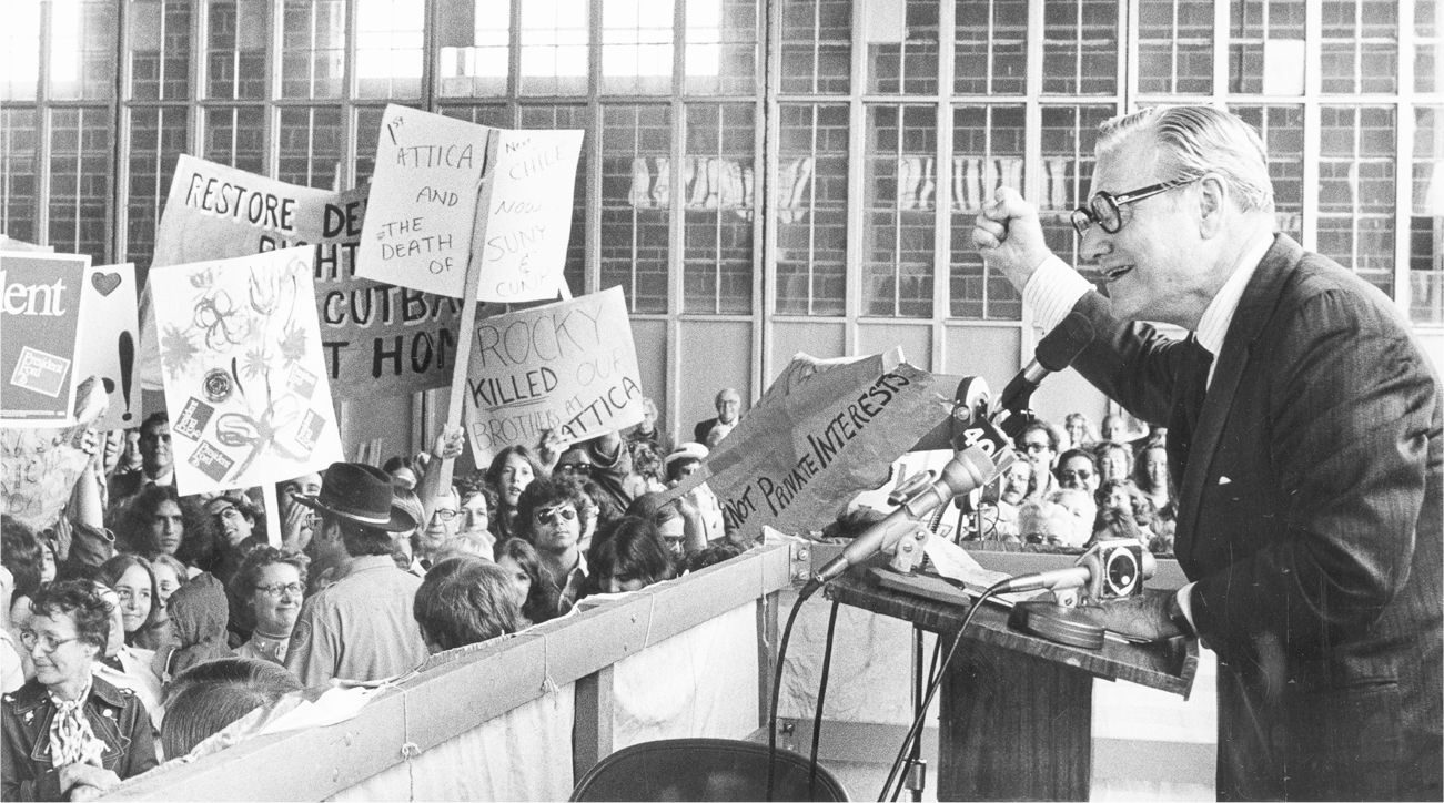 Rockefeller gives a speech from a podium with his fist raised in the air for emphasis. He speaks into a microphone while people in the audience hold signs that express support for and opposition to Rockefeller and the 1976 Republican presidential campaign. Two of the most prominent signs include the text “1st Attica Now Chile and Now the Death of SUNY & CUNY” and “Rocky Killed Our Brothers at Attica.”