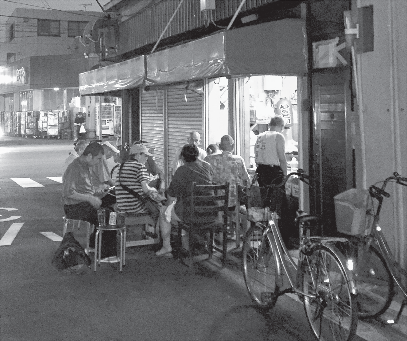 As night falls on the streets behind them, a small group of men sit before a hole-in-the-wall establishment with yellow lighting. They sit on stools, and one man has his head slightly lowered, as if to reminisce.