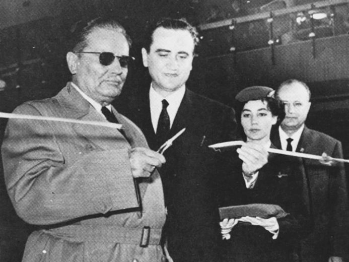 Figure 8.1. A man in a trench coat and wearing sunglasses cuts a ribbon with small scissors, marking the opening of Belgrade’s new airport. A JAT stewardess stands by.