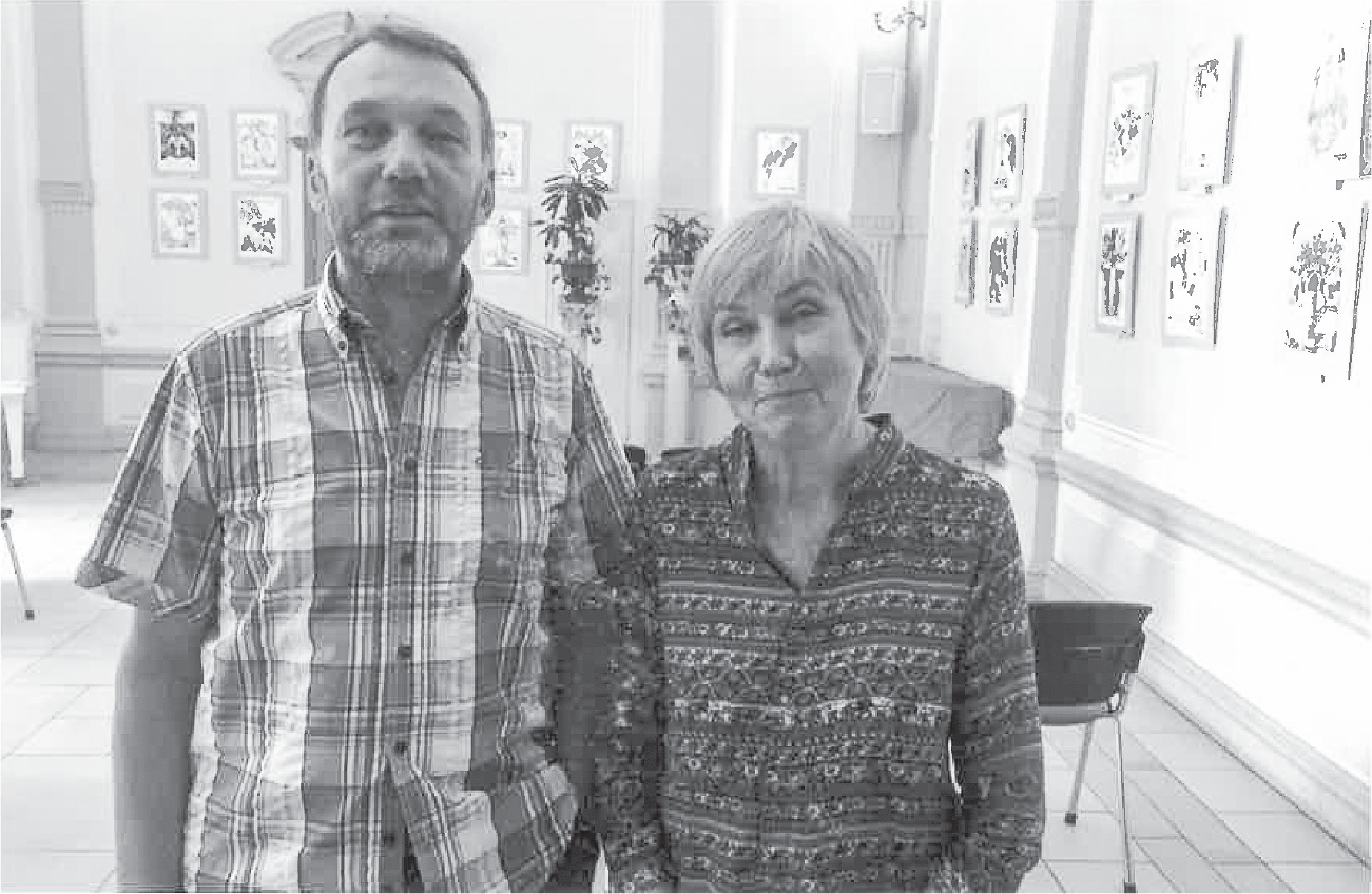A middle-aged couple, smiling for the camera, stand in a room with art on the walls arranged in squares of four pictures each.