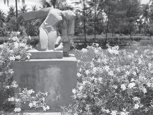 A large stone statue depicts a woman collapsing as she is shot dead, dropping her infant child at her side. The statue is surrounded by flowers on a sunny day at the My Lai Memorial in Vietnam.