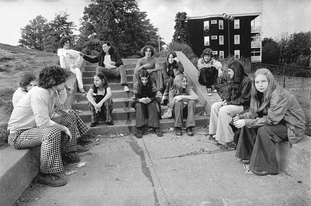 Figure 40. A group of young teenagers sit on cement steps in a park. All of them are wearing bell-bottoms. The boys have long hair, as do the girls. They are chatting with each other. In the back one boy tugs another’s shirt.