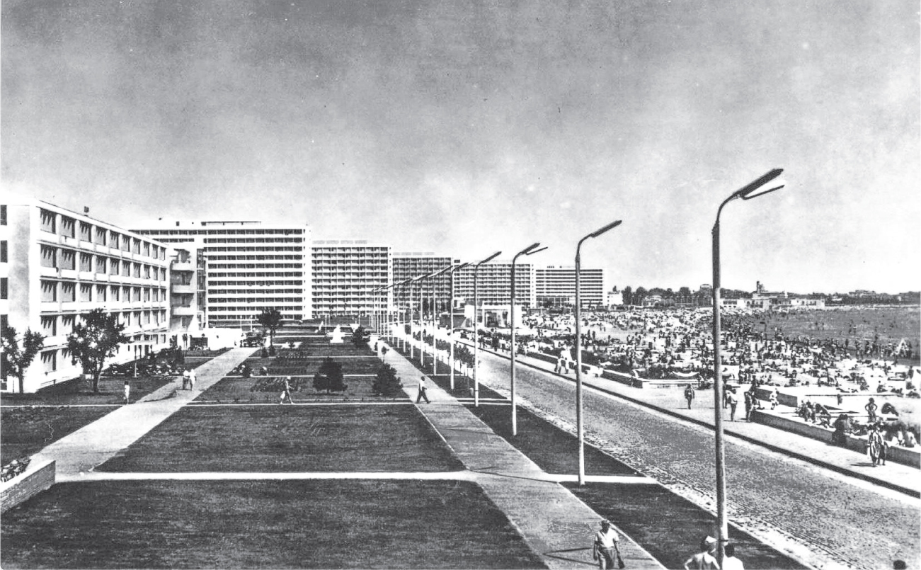 A long-distance photo of five hotel buildings with a plaza and walkways in front of them. On the far right of the picture is the beach, crowded with people.