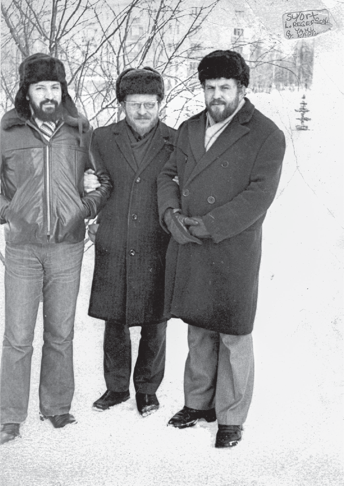 Three bearded men in fur hats stand in the snow with thin tree branches behind them. The center man wears glasses and holds the arms of the other two.