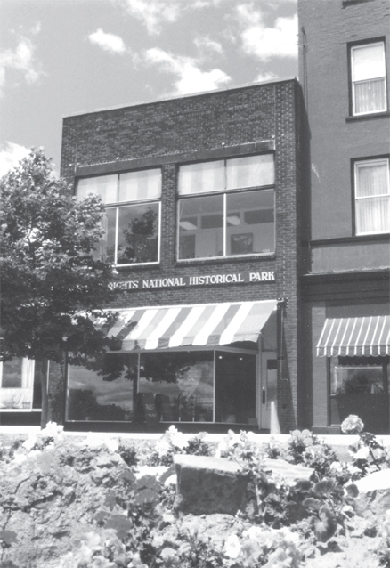 Figure 8. A two-story brick commercial building has a plate-glass storefront with a full-width striped awning and a pair of large plate-glass second-floor windows. A simple sign extending over the awning reads “Women’s Rights National Historic Park.”