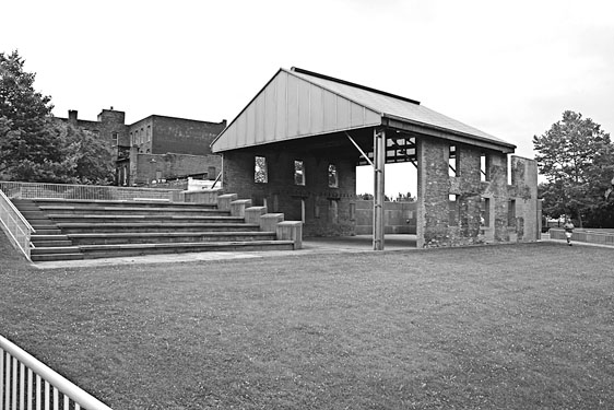 Figure 20 More distant view of the winning design shows the open structure’s two plain brick walls with their sheltering roof, plus an adjoining six-tier concrete amphitheater. A slightly downward-sloping grass lawn is in the foreground.