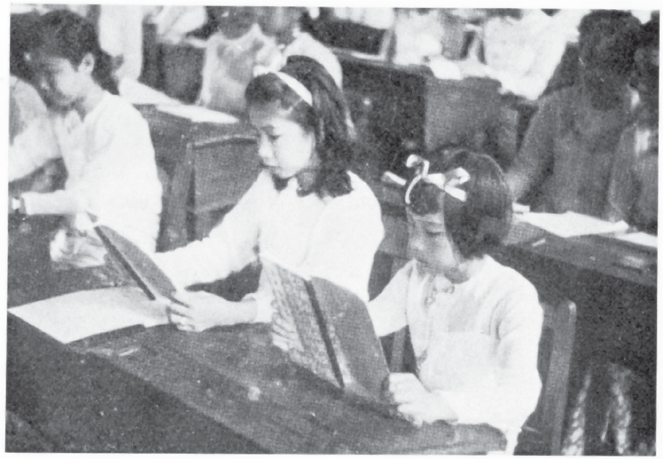 Auntie Amy and one of her sisters, both wearing a white Burmese blouse and a ribbon headband, sit next to one another at a desk and stare intently at what appear to be Japanese textbooks.