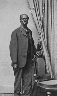 A long-faced, balding man stands with his hand on the back of a chair. He is dressed in a full-cut, three-button wool suit and square-cut shoes.