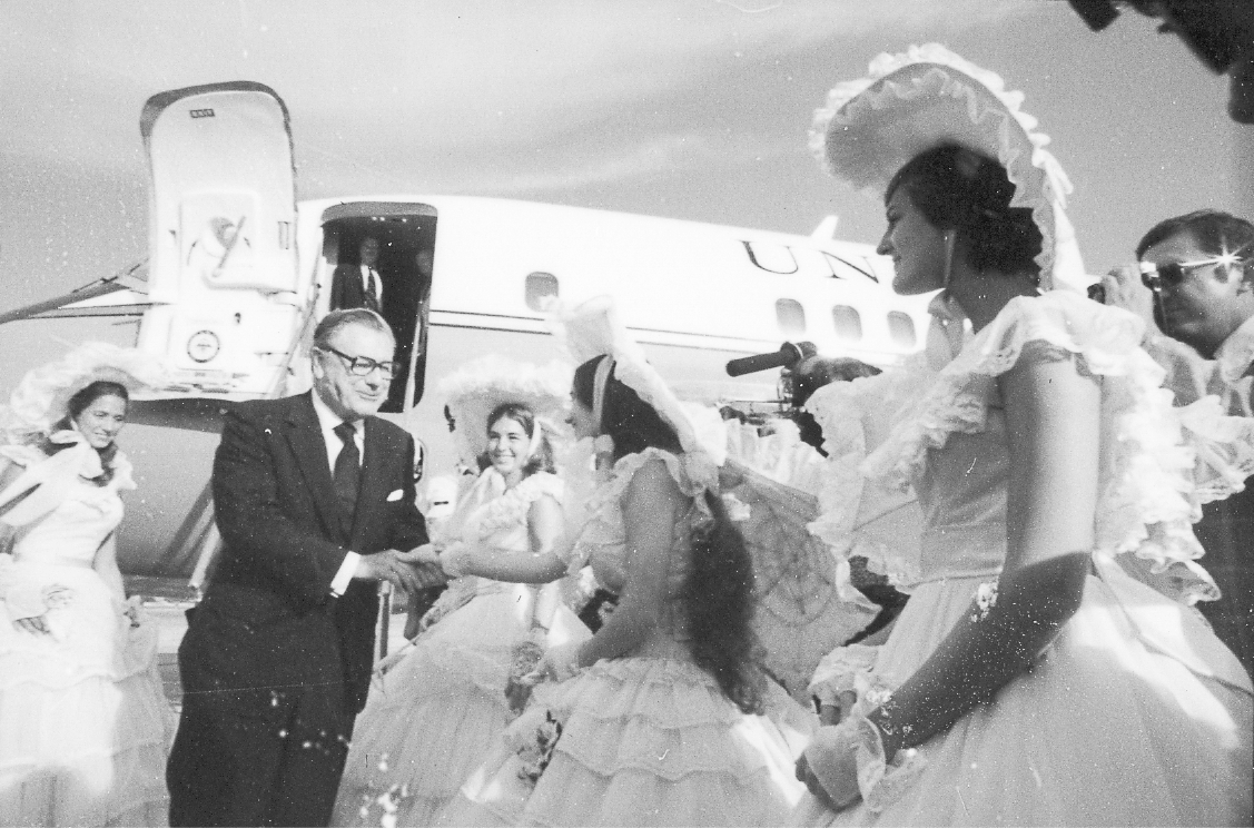 Rockefeller deplanes an Air Force Two plane. At the bottom of the stairs, he shakes the hand of a woman wearing a re-creation of an antebellum dress accessorized with a large hat and parasol. There are four women in similar costumes, while members of the press observe.