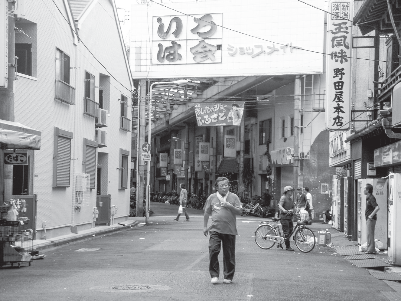 With a towel wrapped around his neck, a man walks away from the entryway to an arcade, where a <i>Tomorrow’s</i> Joe anime banner swings beneath the overhang. Off to the right side of the photo are vending machines, an overflowing garbage can, and more men, standing aimlessly or walking about.